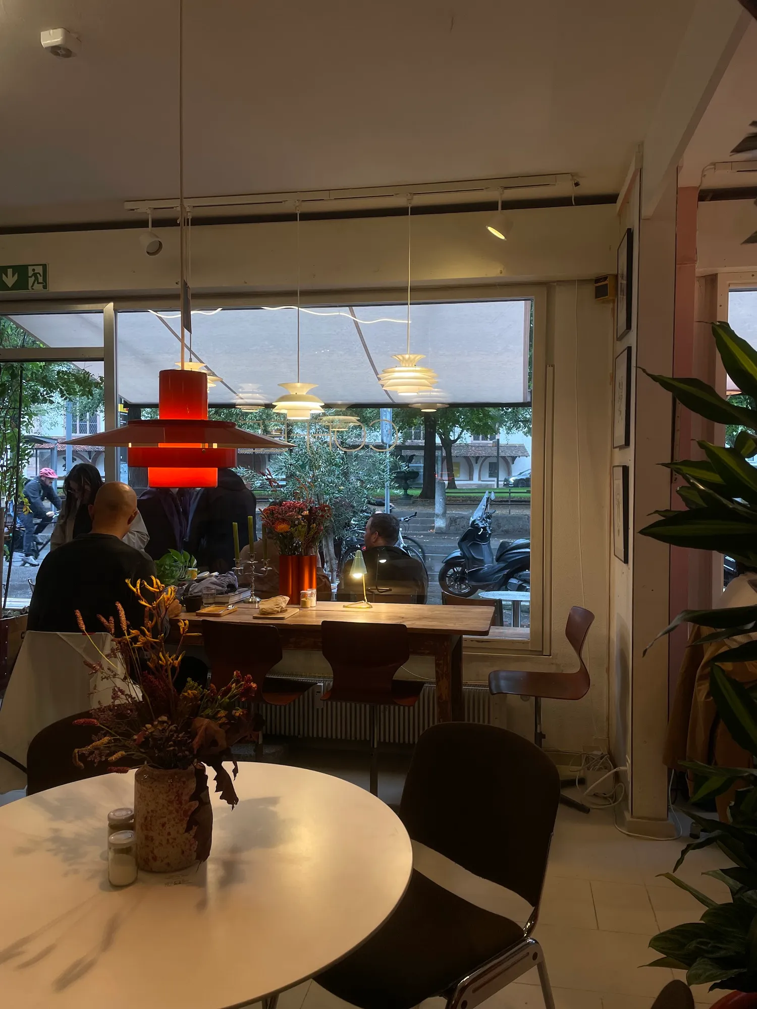 Innenraum mit oranger Pendelleuchte, runden Tischen, Trockenblumen und grossem Fenster zur Strasse