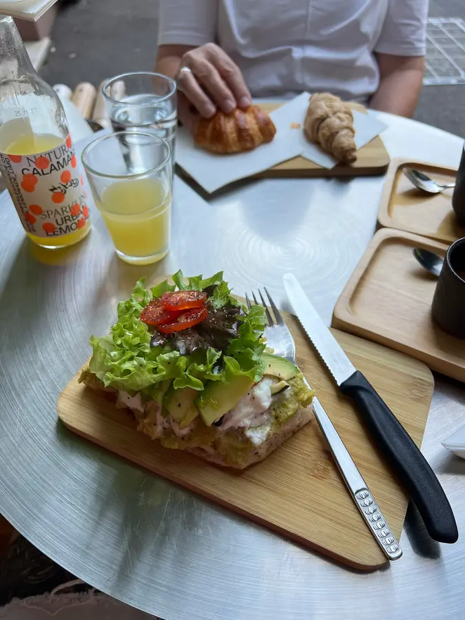 Belegte Focaccia mit Avocado und Salat, dahinter Croissants und Limonade