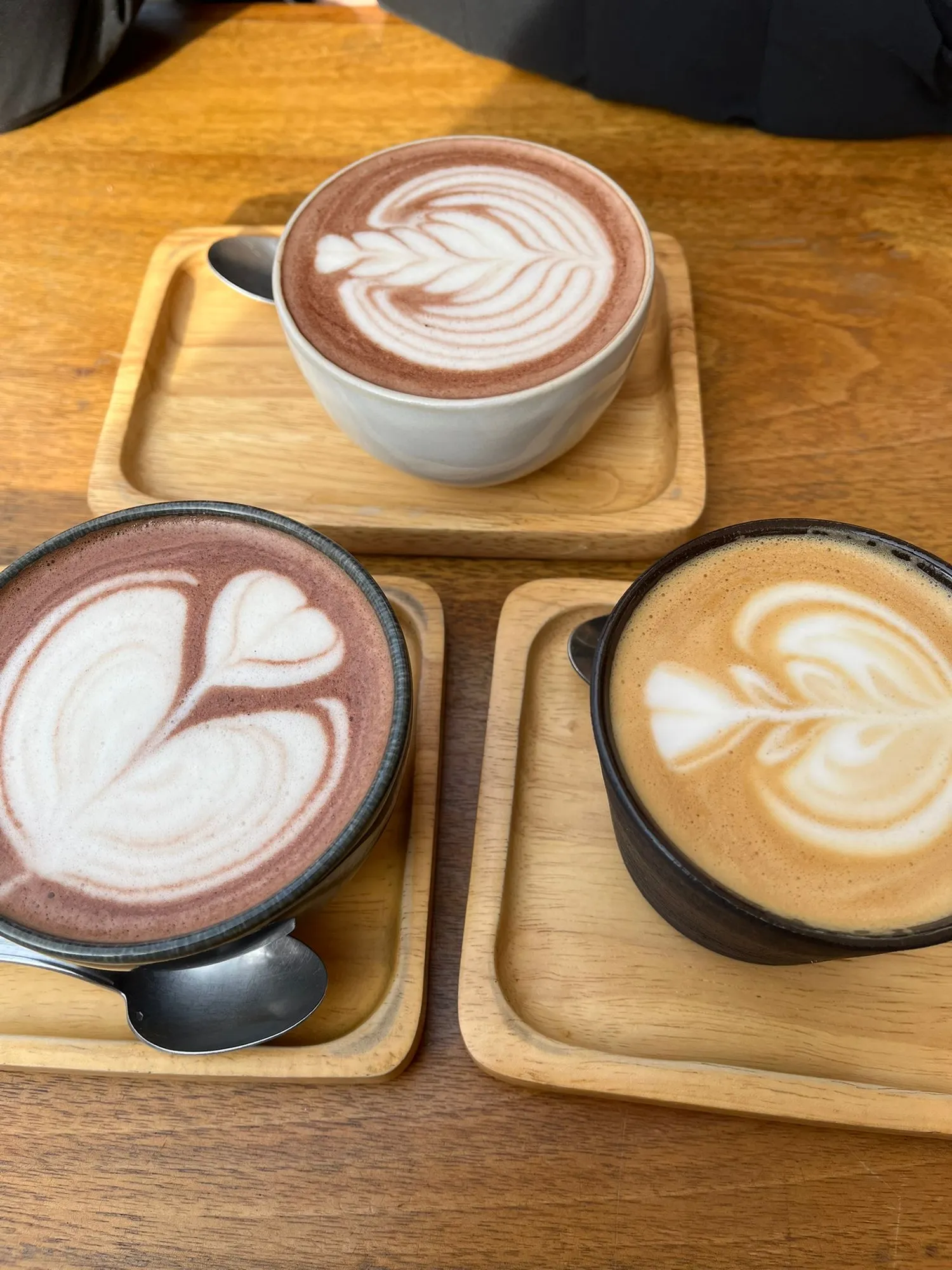 Drei Tassen mit Latte-Art auf Bambus-Tabletts: zwei heisse Schokoladen und ein Cappuccino